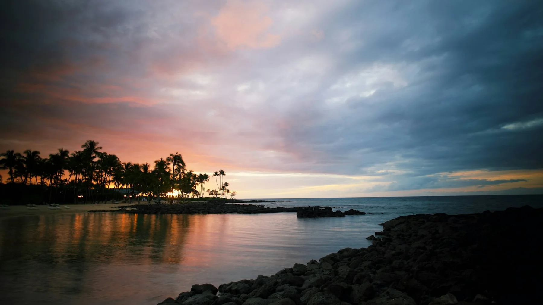Palm trees silhouetted against a pink and orange sunset over a calm bay on Hawaiʻi's Big Island